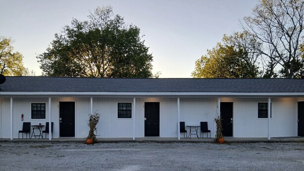 A motel with a row of rooms, each with a door and a window. There are two tables and chairs outside each room.