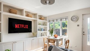 Un soggiorno moderno con televisione a schermo piatto, mobili bianchi, una zona pranzo con tavolo e sedie, e un'ampia finestra con vista su un'area verde.