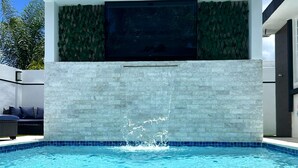 A modern outdoor pool with a waterfall feature, a lounge area with a black mat, and a glass of water with a lemon slice. - Outdoor Pool with Bar-B-Q & Television, Indoor Pool Table, Indoor Bar, PS5, Etc. (Luquillo)