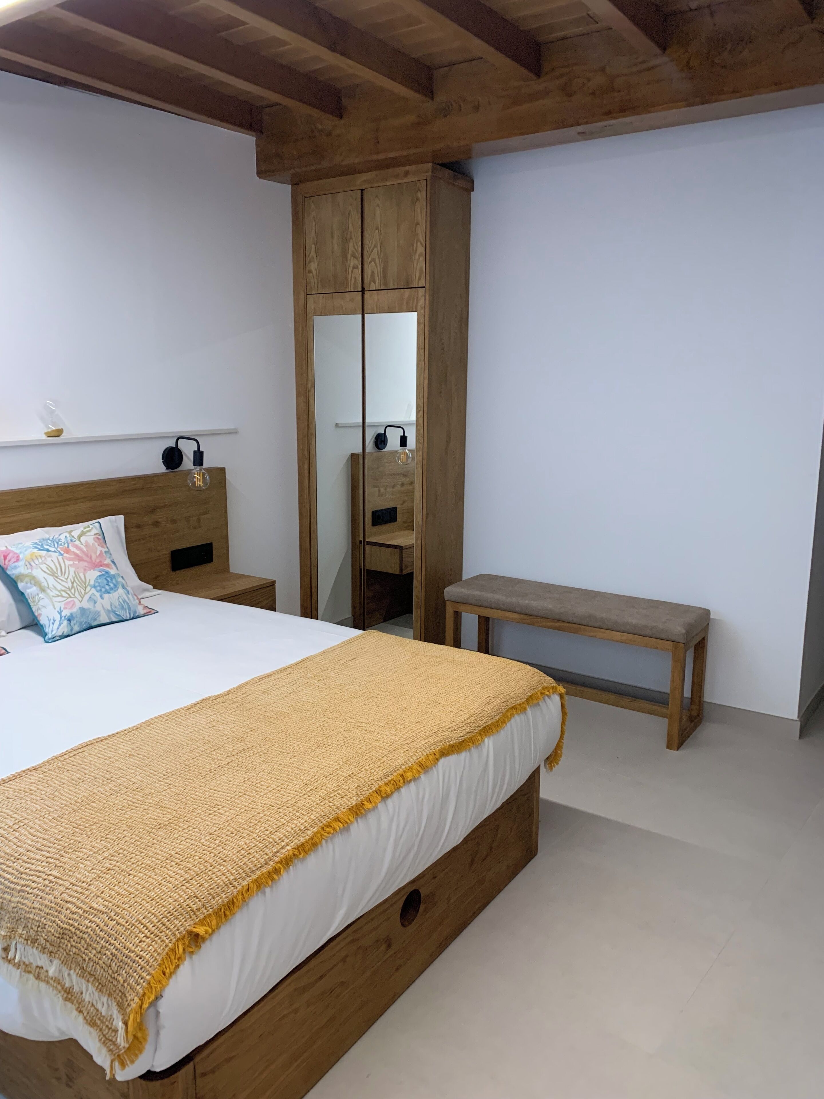 Quarto com estrutura de cama de madeira, um banco, guarda-roupa e espelho.