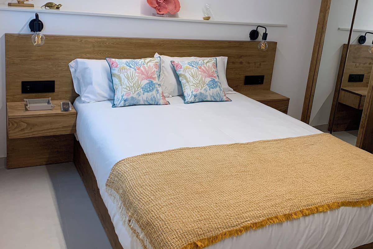 A modern bedroom with a wooden headboard, a white bedspread, and a yellow throw blanket. There is a nightstand with a lamp and a small decorative item on the shelf above the bed.