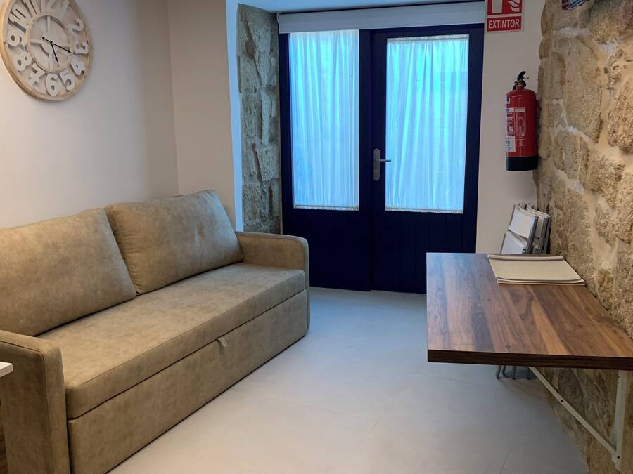 Una habitación con sofá color beige, una mesa de madera, pared de piedra, extintor y una puerta con cortinas.