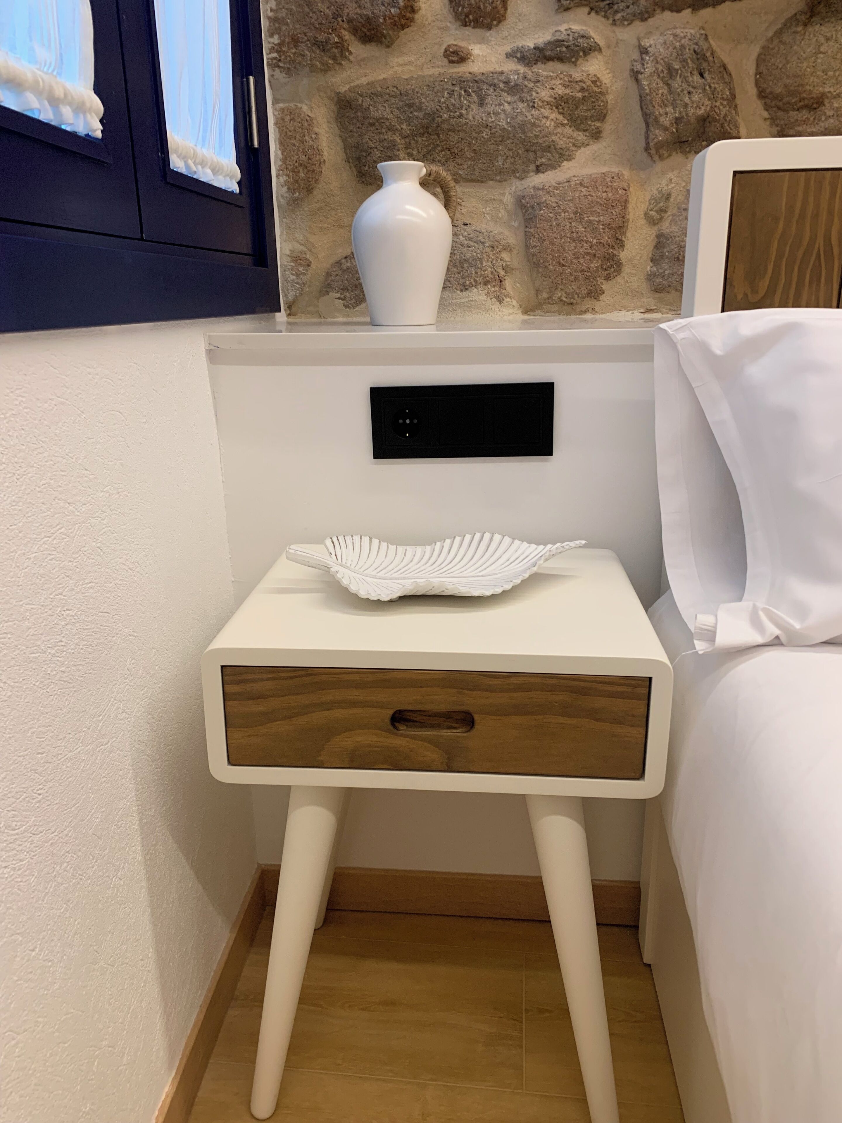 Una mesita de noche pequeña con un jarrón blanco y un objeto decorativo encima, un cajón de madera y un enchufe en la pared.