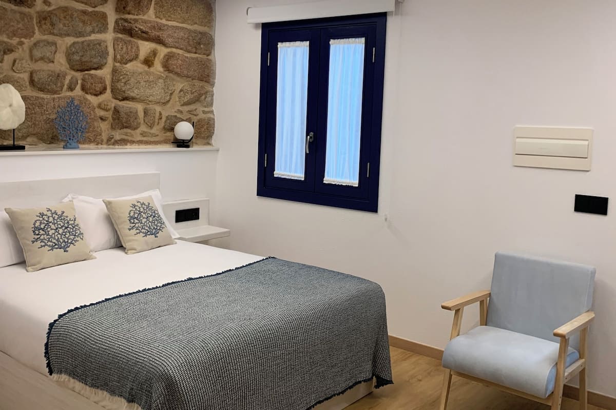 A bedroom with a stone wall, a bed with a grey blanket, a chair, and a window with curtains.