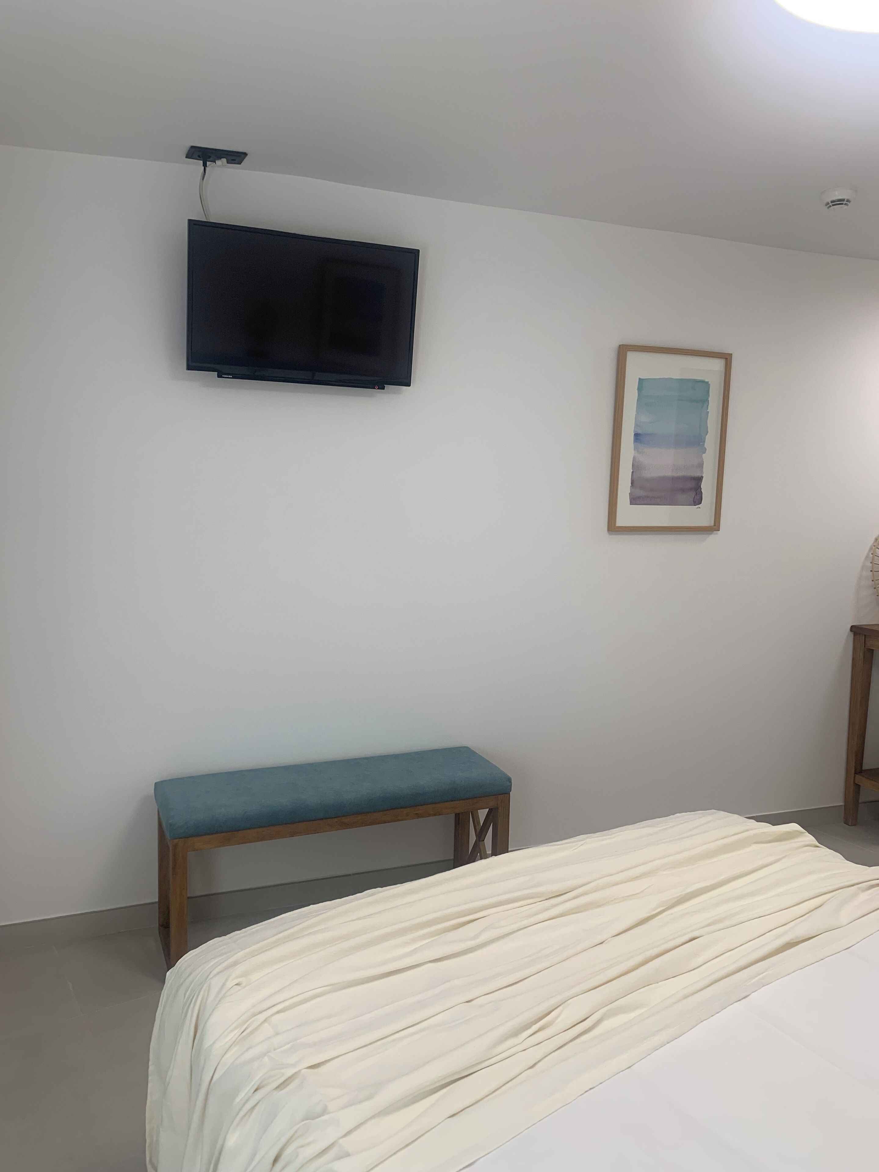 Une chambre à coucher avec un lit, un banc, un téléviseur fixé au mur et un tableau encadré.