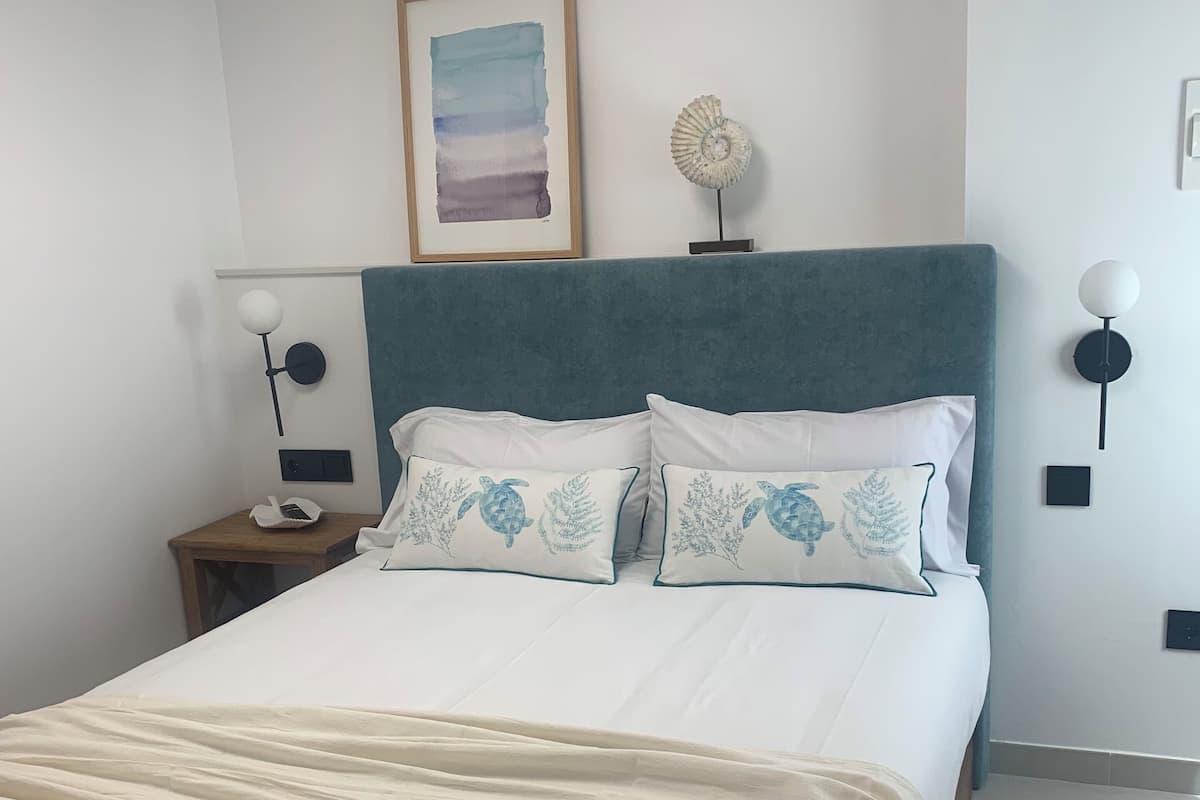 A neatly made bed with white linens and blue floral pillows, a teal upholstered headboard, a framed artwork above the bed, and two wall-mounted lamps on either side.