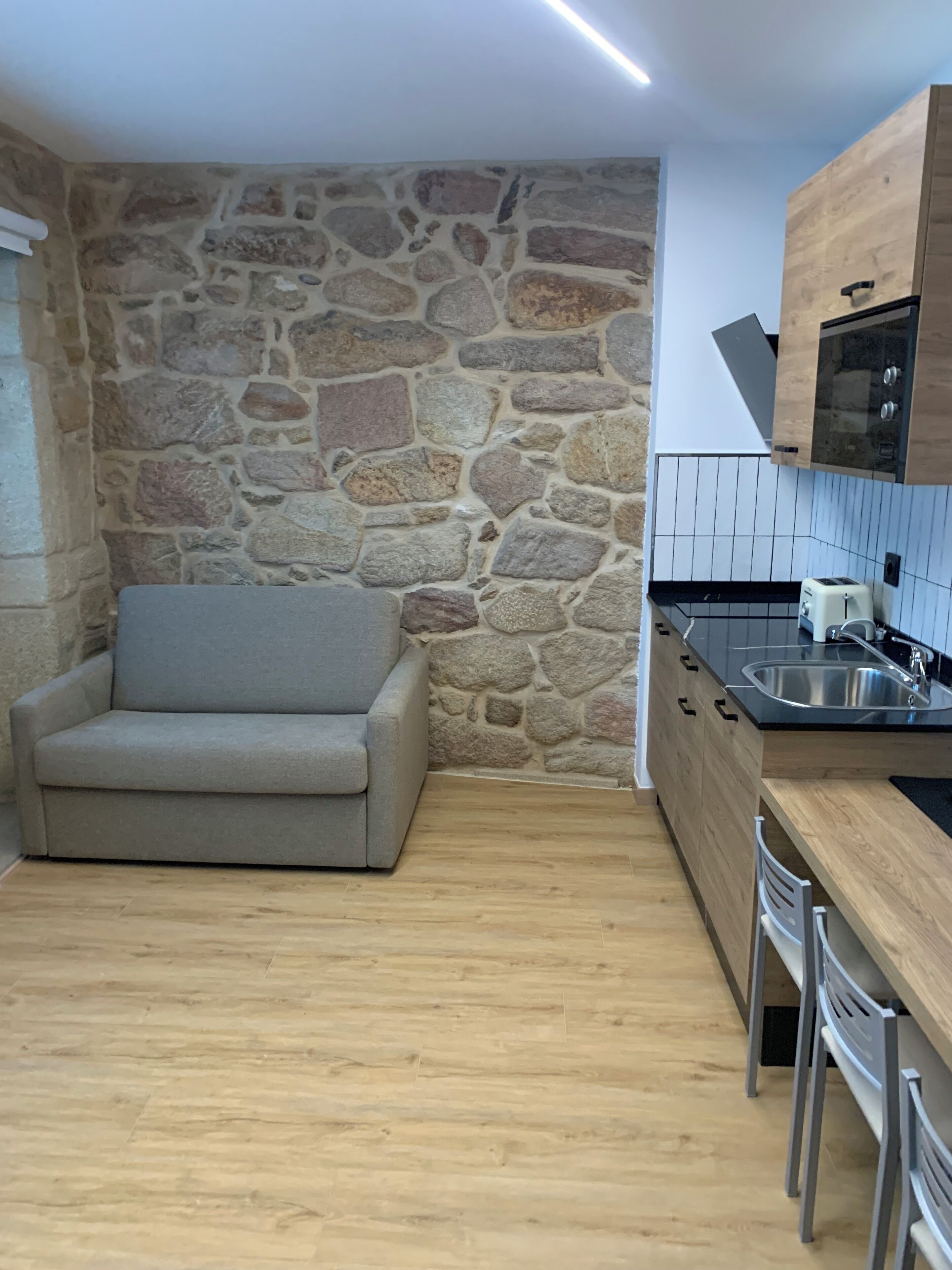 Una cocina compacta con pared de piedra, armarios de madera, sofá gris y salpicadero de azulejos.
