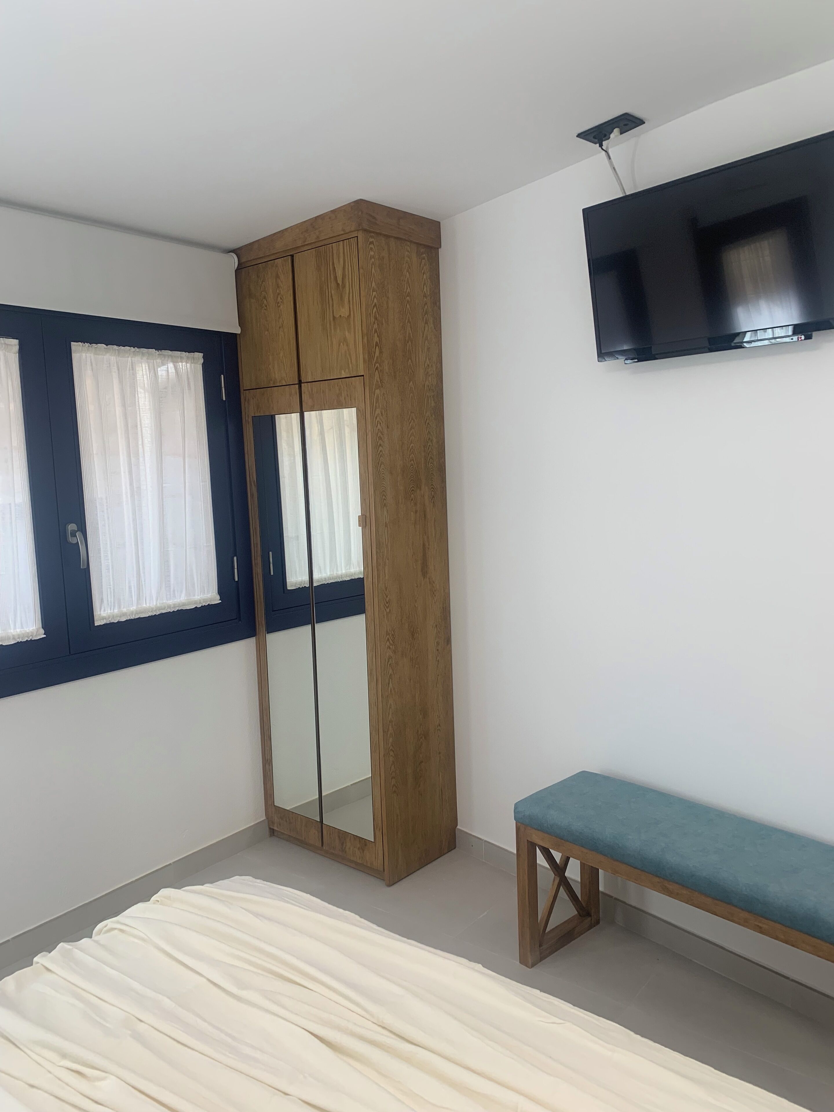 Un dormitorio con un armario, un banco, un televisor montado en la pared y una ventana con cortinas.