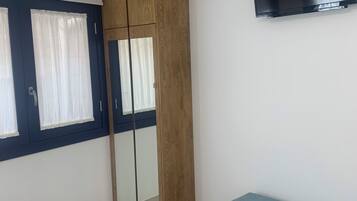 Un dormitorio con un armario, un banco, un televisor montado en la pared y una ventana con cortinas.