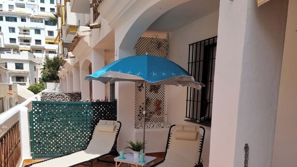 Un balcone con due poltrone da relax e un ombrellone blu, pavimento piastrellato e vista sulla città.