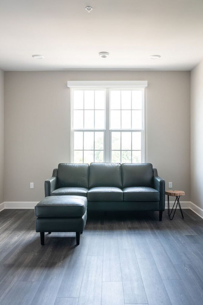 Un salón espacioso con un sofá seccional de cuero oscuro y un reposapiés, una mesita redonda y un ventanal que proporciona luz natural.