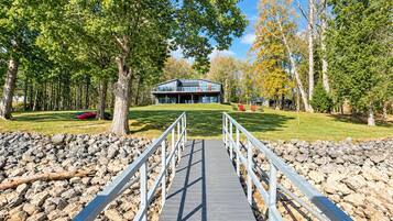 A wooden bridge leading to a modern building with a glass facade, surrounded by trees and a grassy area.