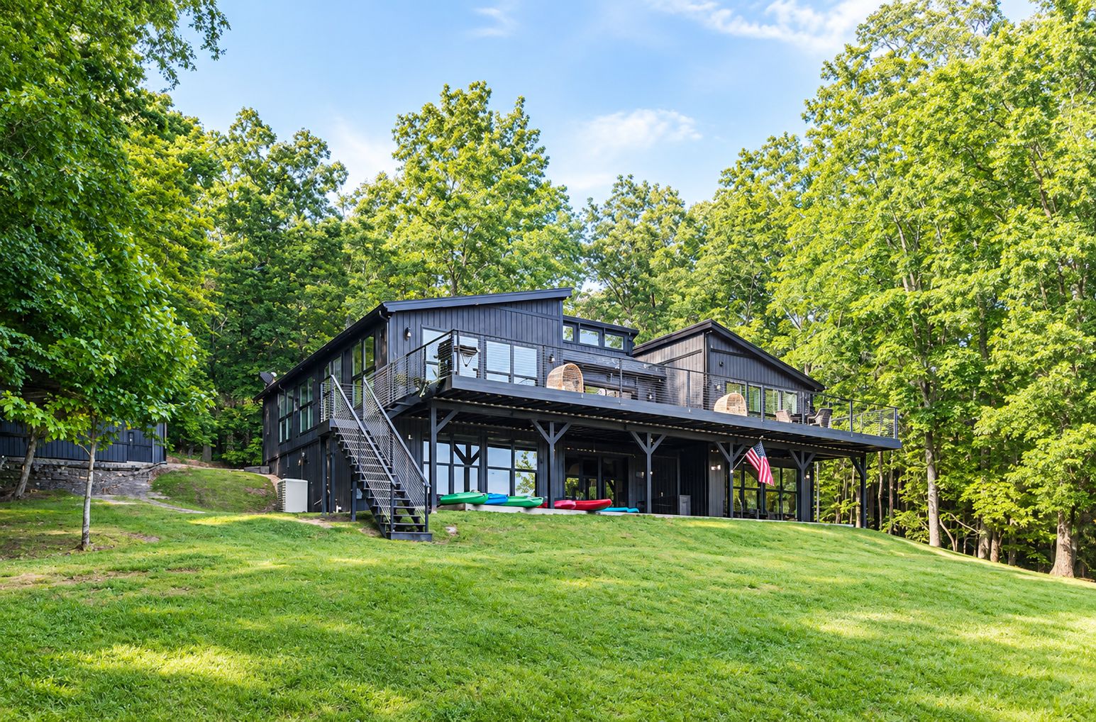 A modern two-story house with a large deck, surrounded by lush greenery.