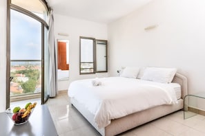 A hotel room with a large bed, a view of the city through a window, and a table with fruit.