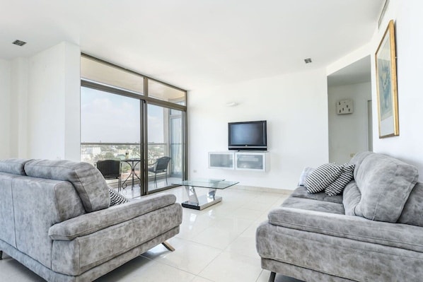 A modern living room with two grey sofas, a glass coffee table, a flat-screen TV mounted on the wall, and a balcony with a glass door.
