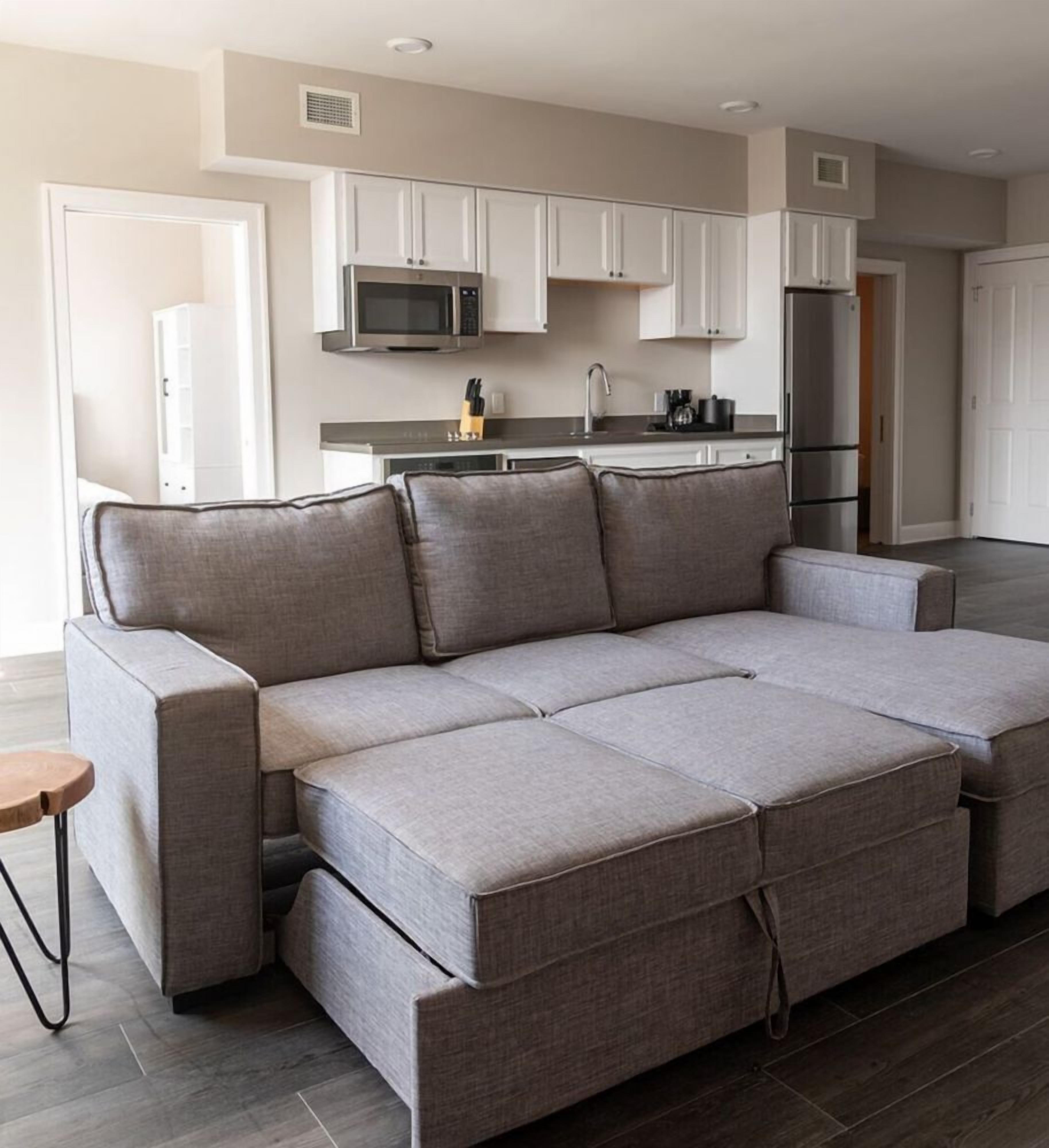 Een moderne woonkamer met een grijze modulaire bank, een klein houten bijzettafeltje, en een keuken met witte kasten, een magnetron en een koelkast.
