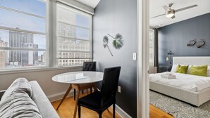 Un dormitorio moderno con cama, una mesa redonda y sillas, vistas de la ciudad a través de amplias ventanas y un ventilador de techo.