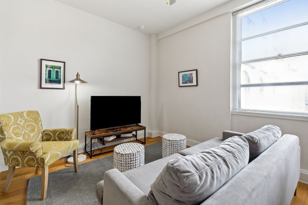 Ein Wohnzimmer mit grauem Sofa, einem gelb gemusterten Sessel, einem Flachbildfernseher auf einem Holzboden, zwei runden Hocker und gerahmten Bildern an den Wänden.