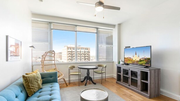 Ein modernes Wohnzimmer mit einem blauen Sofa, einem runden Couchtisch, einem Fernseher auf einer Holzkommode und Blick auf Gebäude durch große Fenster.