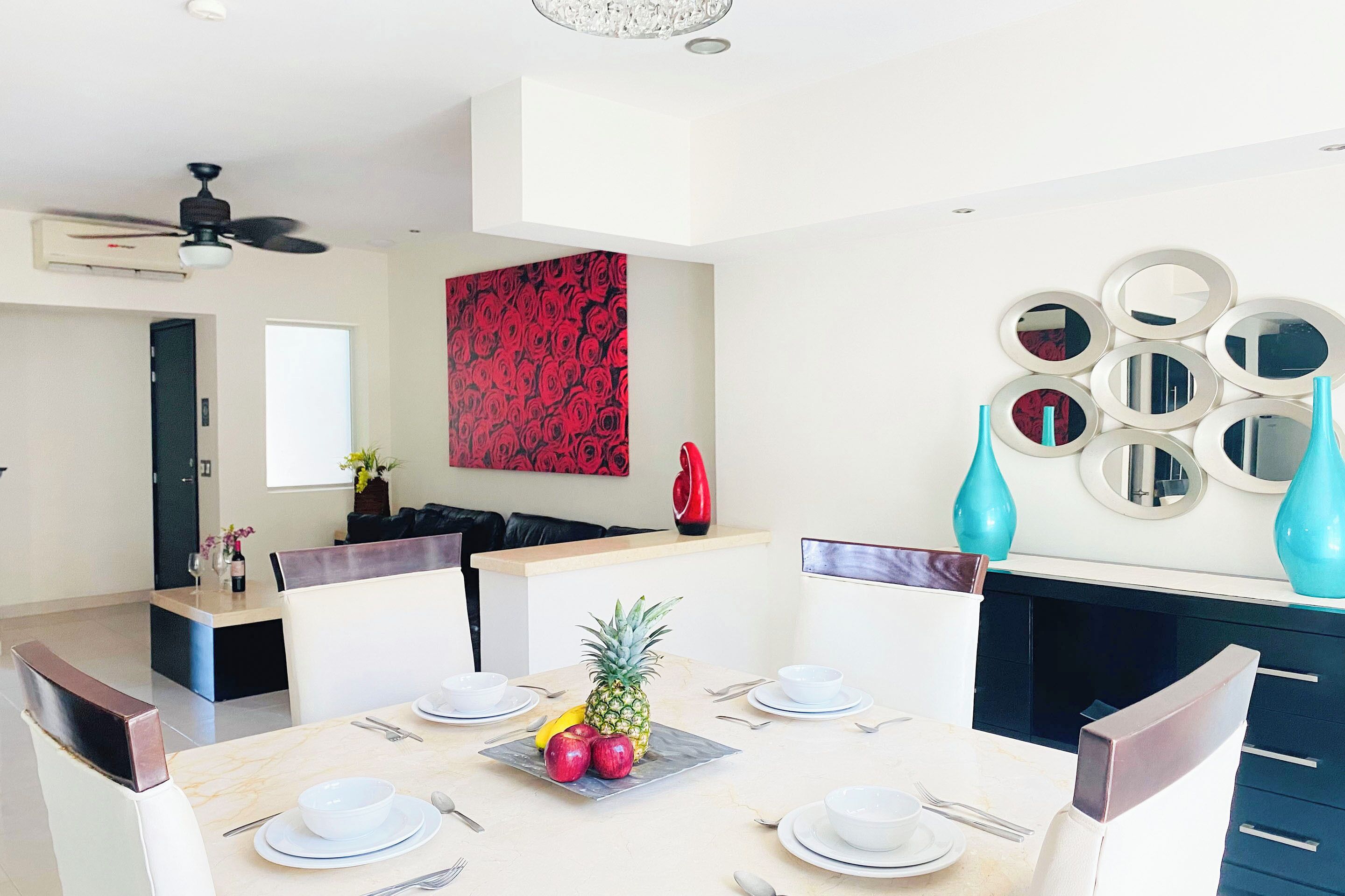 Un comedor moderno con una mesa preparada para cuatro personas, un centro de mesa decorativo con una piña y una obra de arte floral roja en la pared.