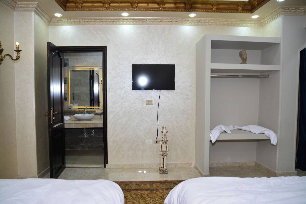 A hotel room with two beds, a TV mounted on the wall, a bathroom with a sink and mirror, and a shelf with a statue.