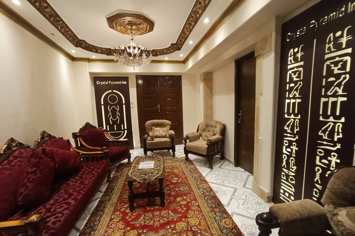 A luxurious hotel lobby with a red patterned sofa, a wooden coffee table, and a chandelier. There is a sign for the Crystal Pyramid Inn and a decorative wall with hieroglyphs.