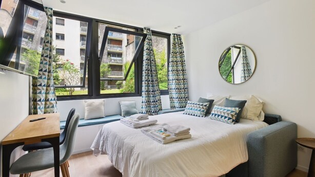 A modern bedroom with a bed, a desk, and a chair. There is a window with curtains, a round mirror on the wall, and a view of buildings outside. - TBS 167 - Refined and bright studio (Paris)