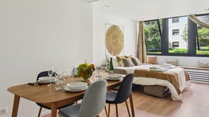 A modern hotel room with a dining area, a bed with a view, and a large window.