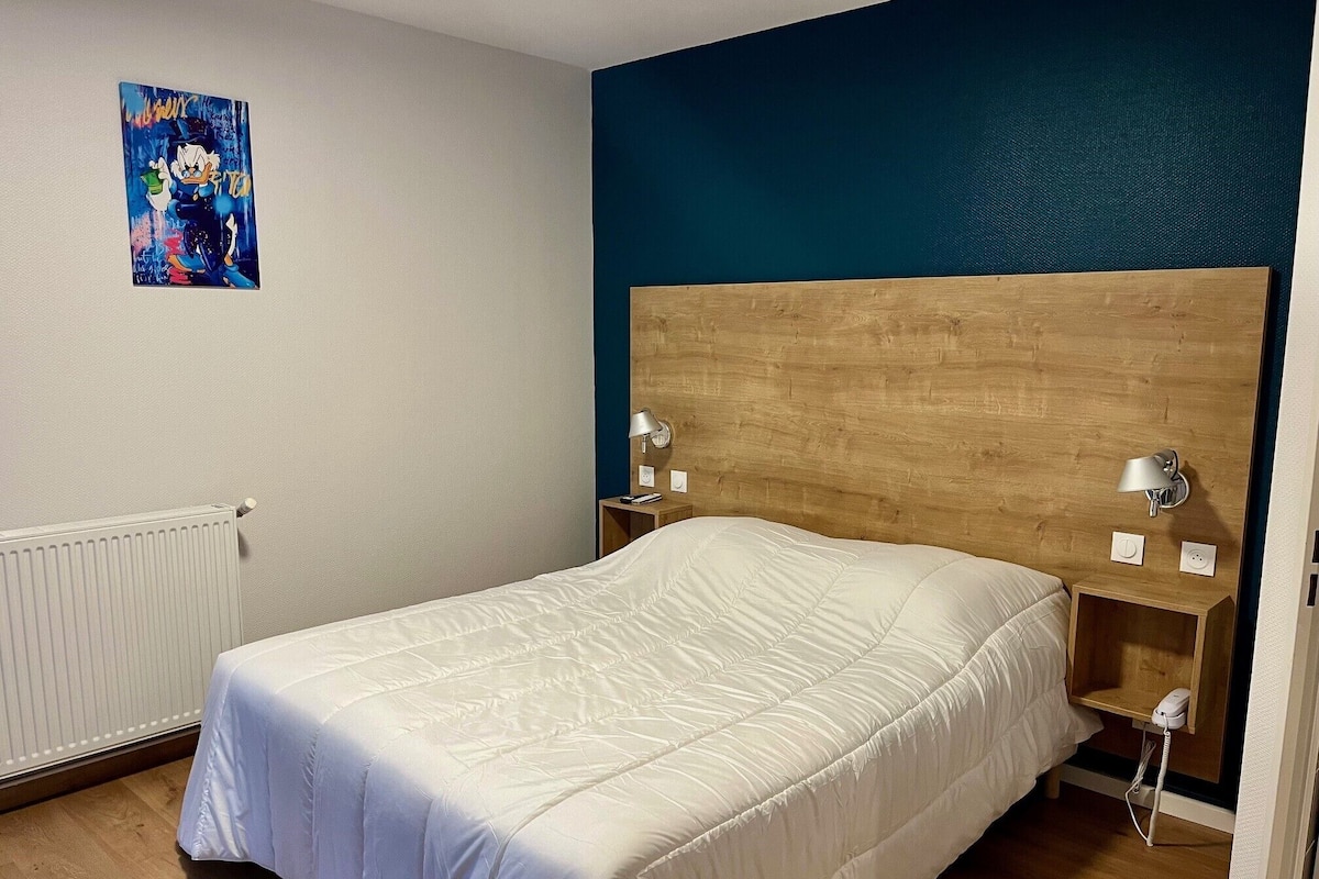 Ein Schlafzimmer mit einem Holz-Kopfteil, einer weißen Bettdecke, einem an der Wand montierten Heizkörper und einem kleinen Nachttisch mit Lampe.
