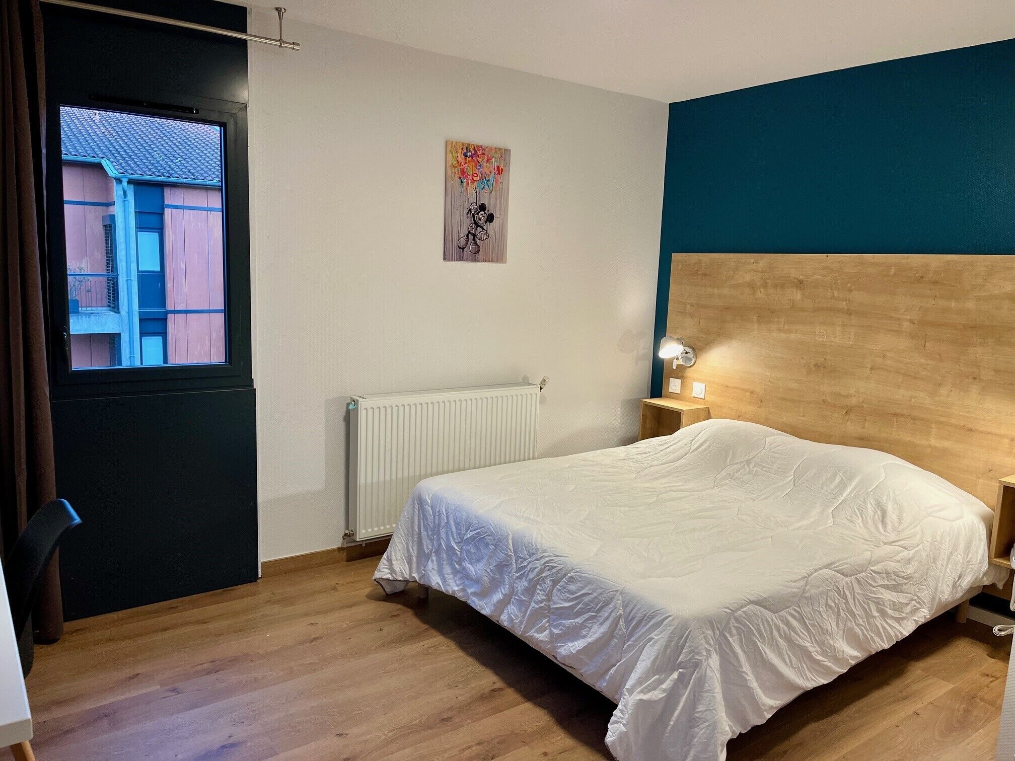 A bedroom with a bed, a window, a radiator, a wooden headboard, and a nightstand with a lamp.