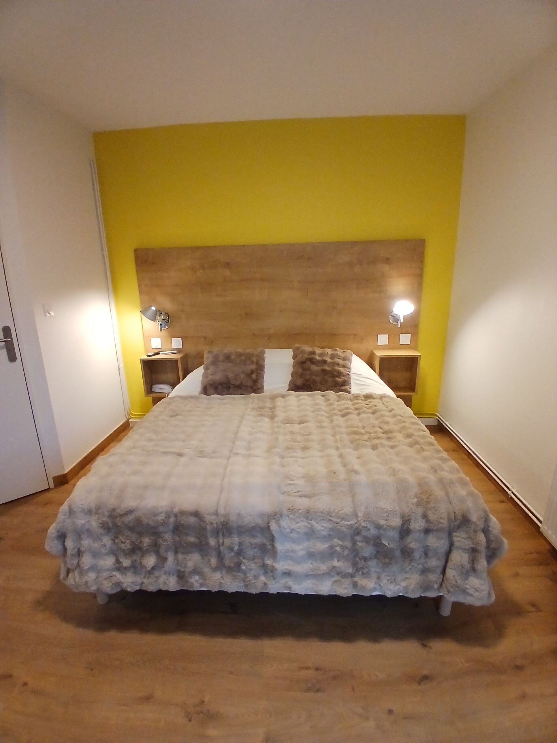 Une chambre à coucher avec une tête de lit en bois, un lit recouvert d’une couette grise et blanche, deux tables de chevet avec des lampes et un mur d’accent jaune.