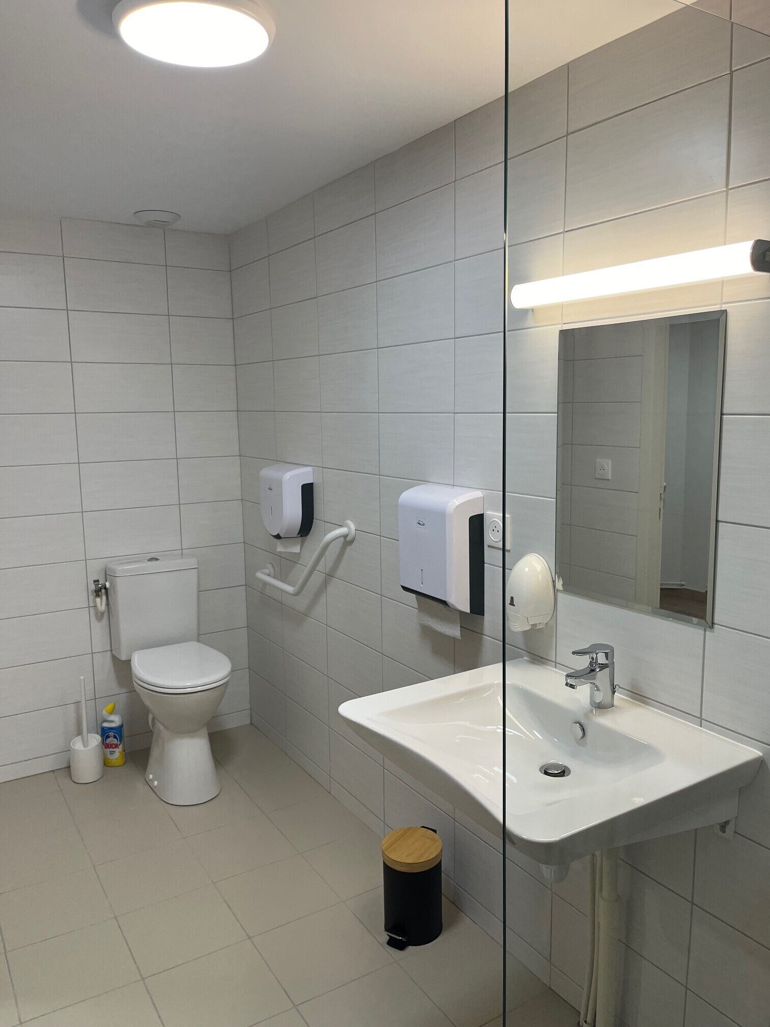 A modern bathroom with a toilet, hand dryer, paper towel dispenser, sink, and a trash bin.