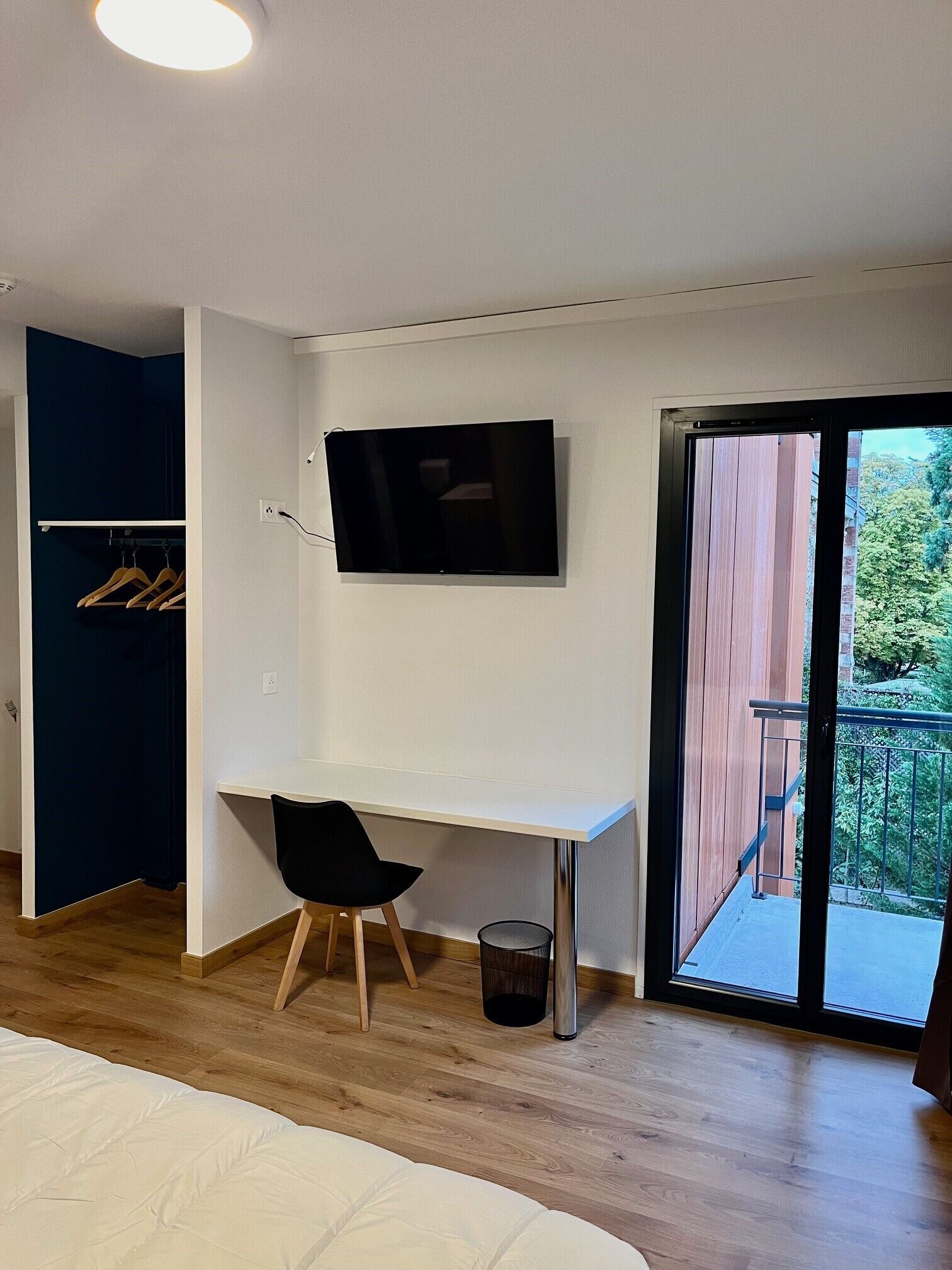 A bedroom with a bed, a desk with a chair, a TV, a wardrobe, and a balcony with a railing.