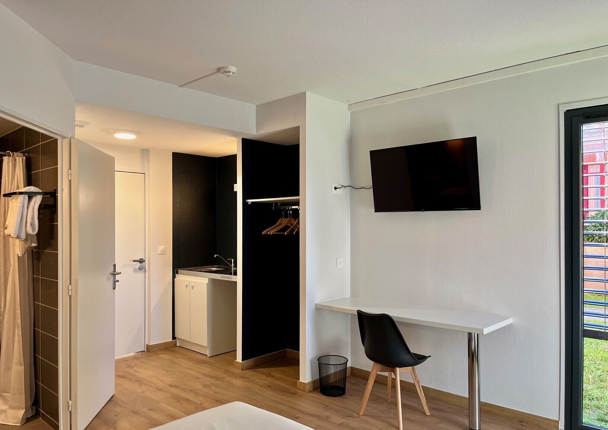 Ein modernes Zimmer mit weißem Schreibtisch, schwarzem Stuhl, Wand-Fernseher und Blick auf ein rotes Gebäude.