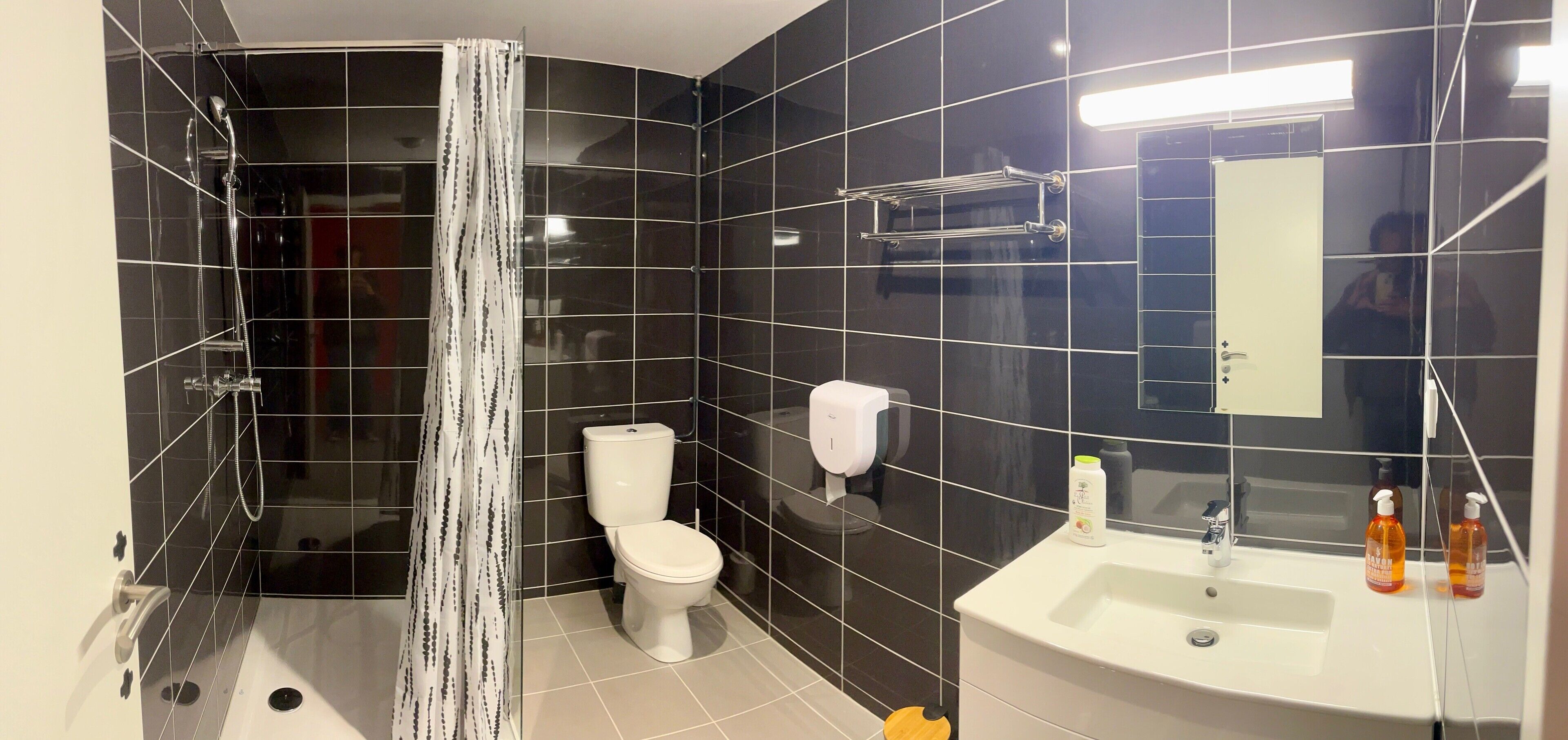 Un bany modern amb rajoles negres, un vàter blanc, una dutxa amb porta de vidre i un lavabo amb dosificadors de sabó.