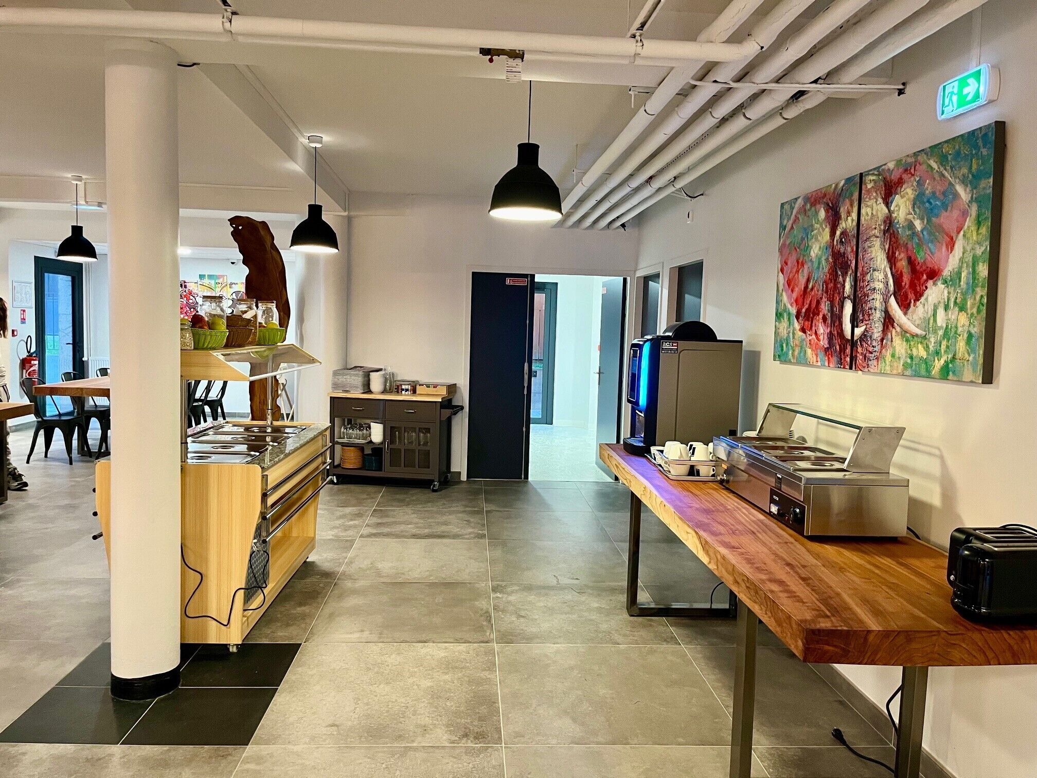 A modern kitchen area with a coffee machine, a toaster, and a wooden table. There is a painting on the wall and a door leading to another room.