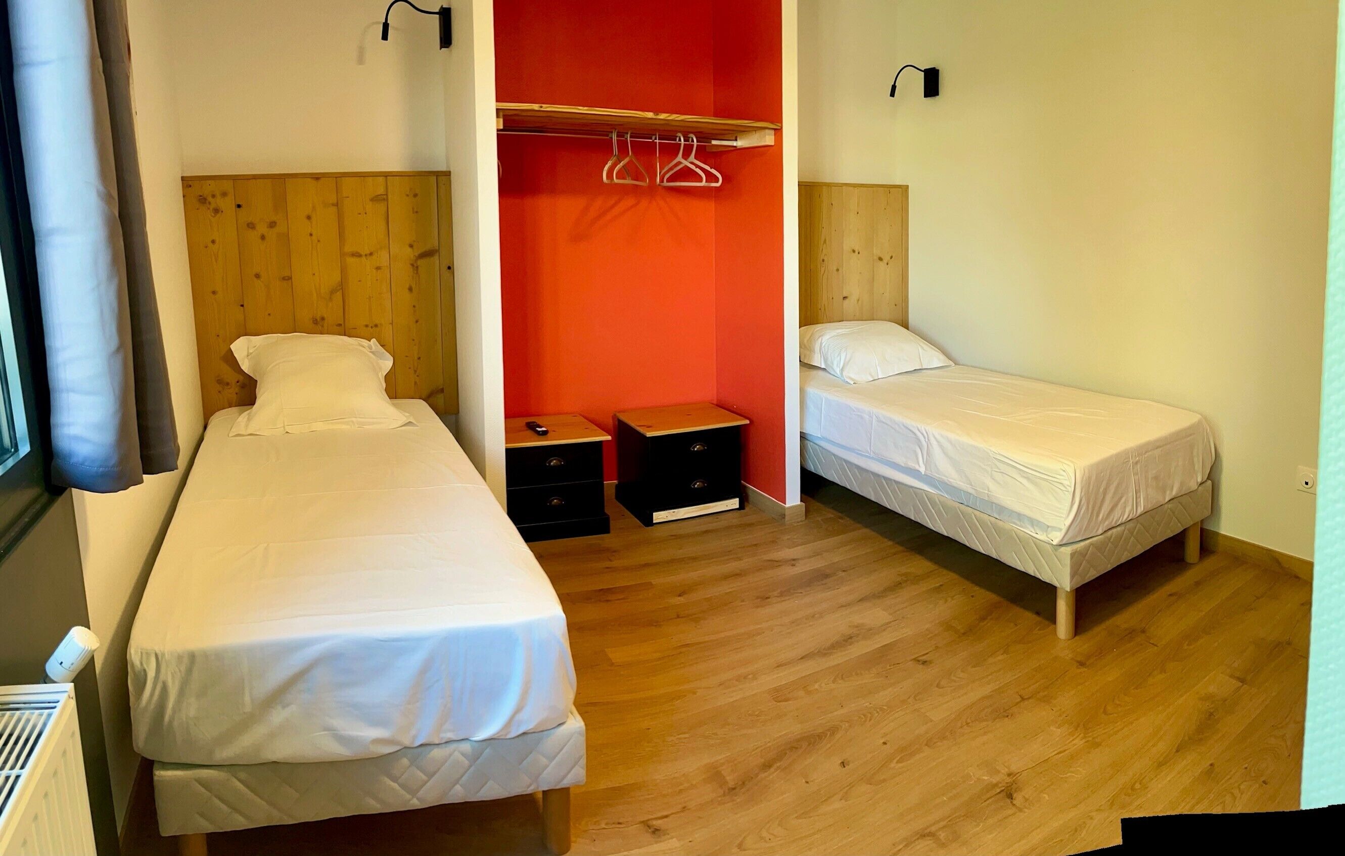 Une chambre avec deux lits, une tête de lit en bois, un mur d’accent rouge et une petite table avec des tiroirs.