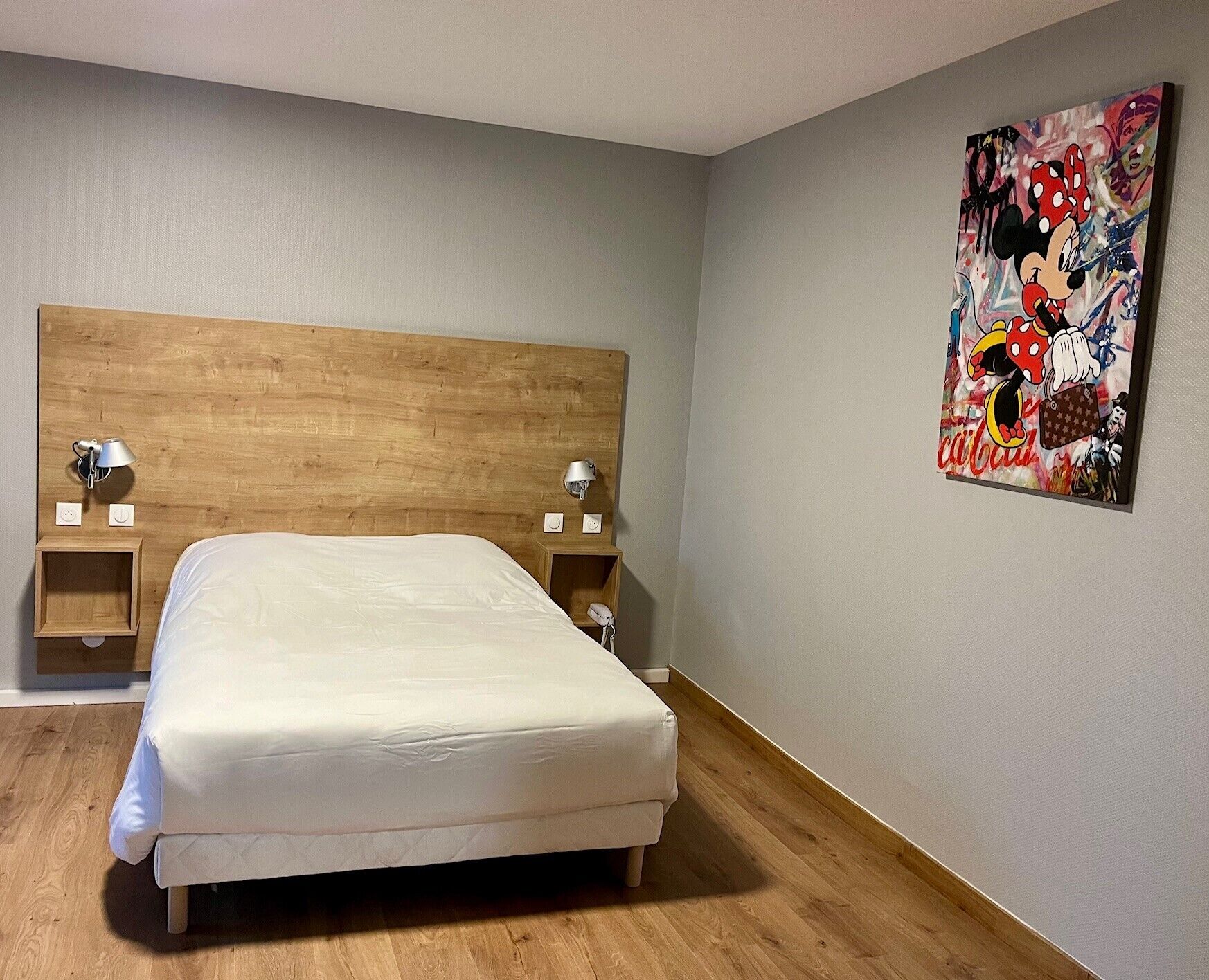 Een slaapkamer met een houten hoofdeinde, een bed met witte beddengoed, twee nachtkastjes met lampen, en een kleurrijk abstract schilderij aan de muur.