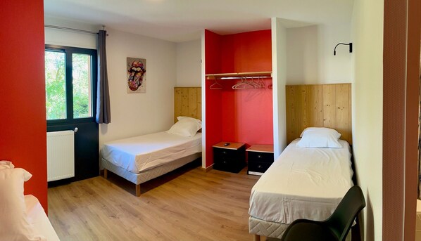 A small, well-lit room with two single beds, a wooden floor, a red accent wall, and a window with curtains. - Les Cimes du Mont Royal (Montrejeau)