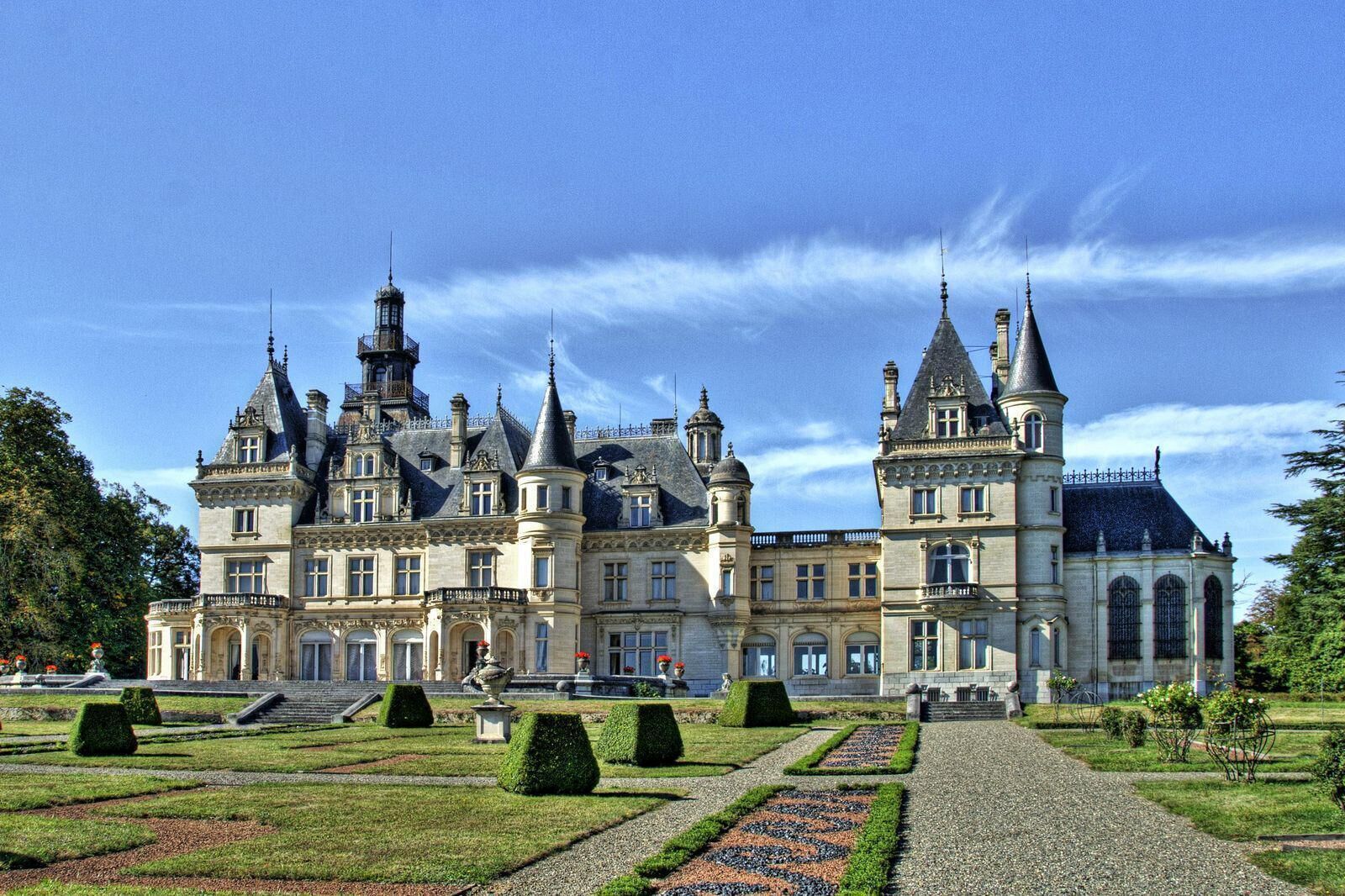 Un edificio grande e histórico con múltiples torres y una fachada simétrica, rodeado de jardines cuidados y un camino de grava.