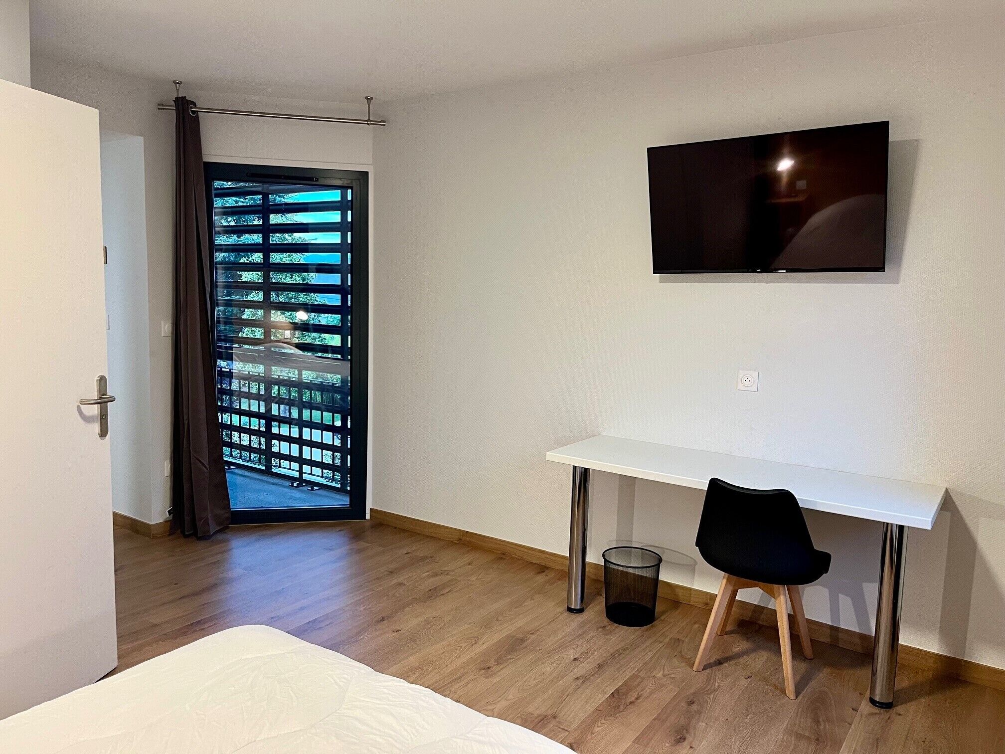 Une chambre d’hôtel avec un bureau blanc, une chaise noire, un téléviseur fixé au mur, une porte donnant sur un paysage verdoyant et une poubelle.