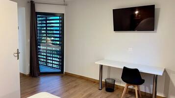 Une chambre d’hôtel avec un bureau blanc, une chaise noire, un téléviseur fixé au mur, une porte donnant sur un paysage verdoyant et une poubelle.