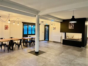 Amplia habitación con una larga mesa de madera y sillas, un sofá negro y una estación de parrilla. Hay ventanas con vistas a edificios, y la habitación tiene un diseño moderno con techos altos a la vista y luces colgantes.