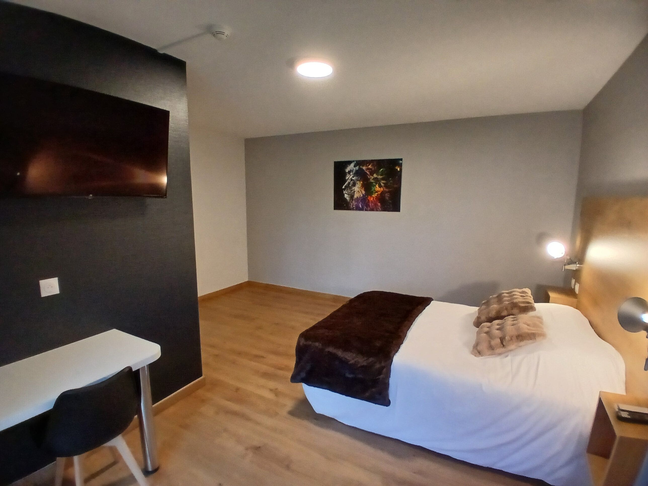 A hotel room with a bed, a desk, a chair, a TV mounted on the wall, and a painting on the wall.