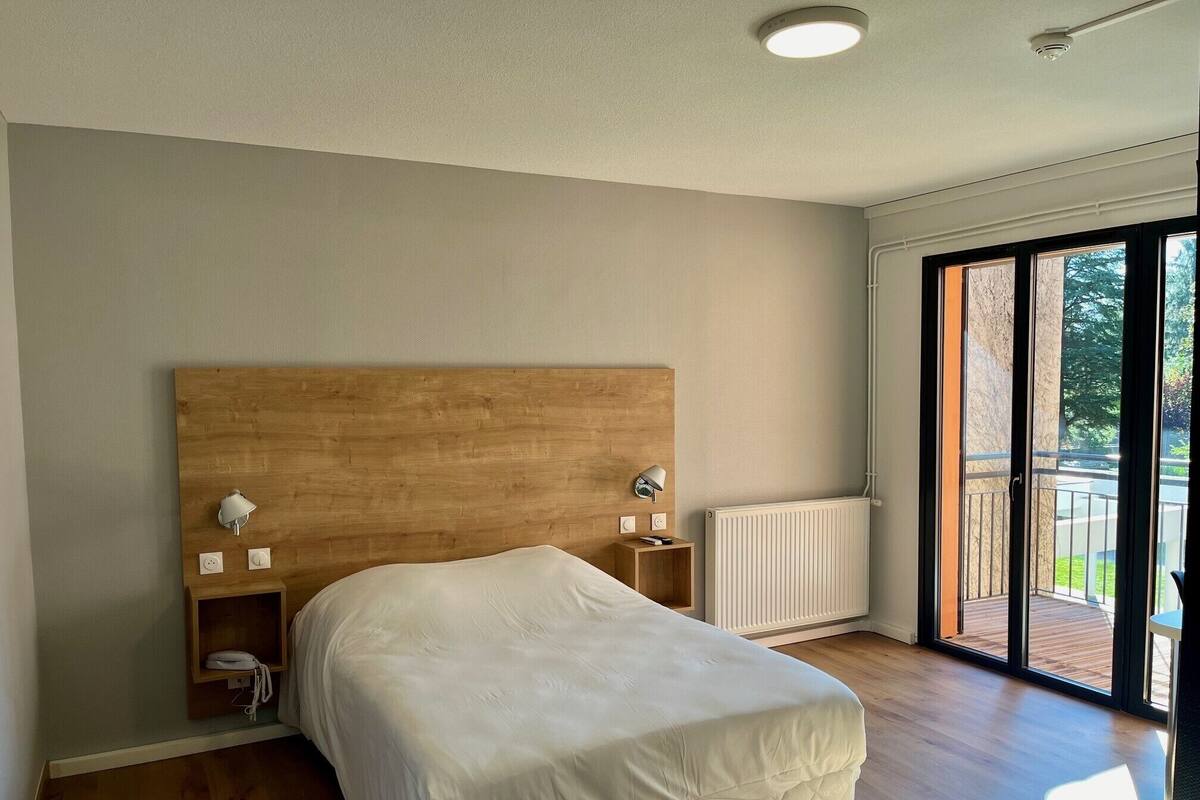 Ein Schlafzimmer mit einem Holz-Kopfteil, weißer Bettdecke, Nachttischlampen, einem kleinen Nachttisch und Blick ins Freie durch eine Glasschiebetür.