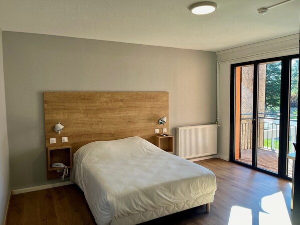 Un dormitorio con cabecera de madera, cubrecama blanco, lámparas de noche, una mesita de noche de madera pequeña y vista al exterior a través de una puerta corrediza de vidrio.