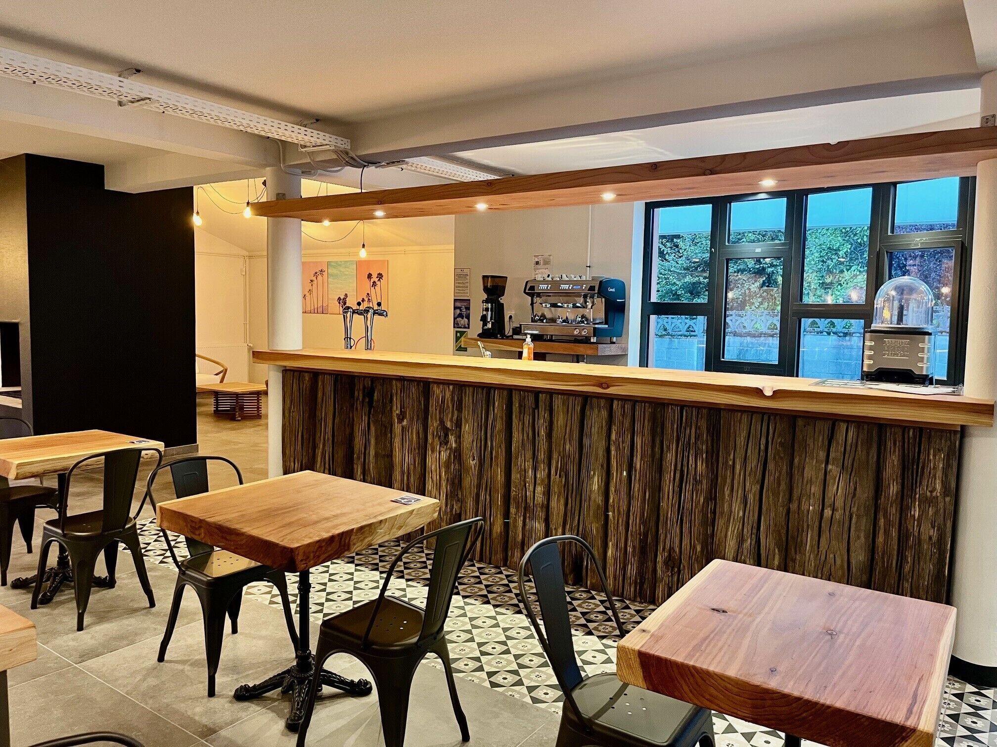 A café with wooden tables and chairs, a bar counter with a coffee machine, and a view of the outdoors through large windows.
