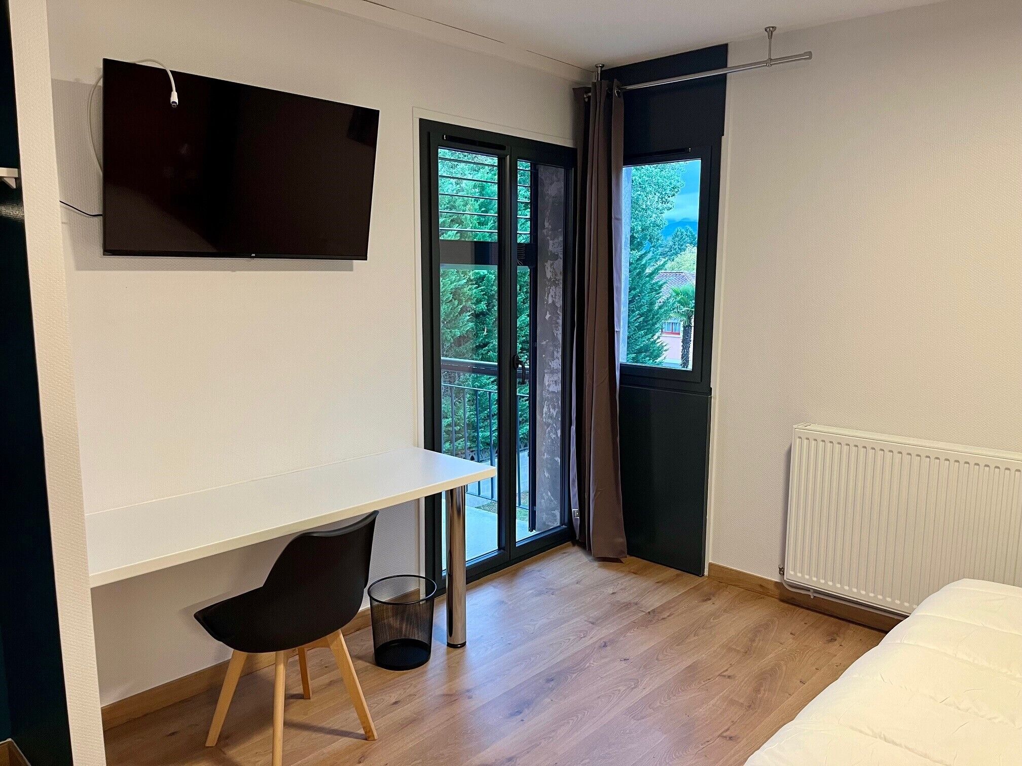 A modern room with a white desk, a black chair, a TV mounted on the wall, a large window with a view of greenery, and a radiator.