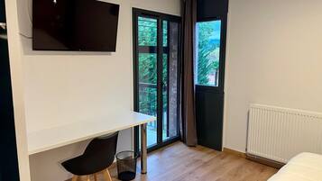 A modern room with a white desk, a black chair, a TV mounted on the wall, a large window with a view of greenery, and a radiator.