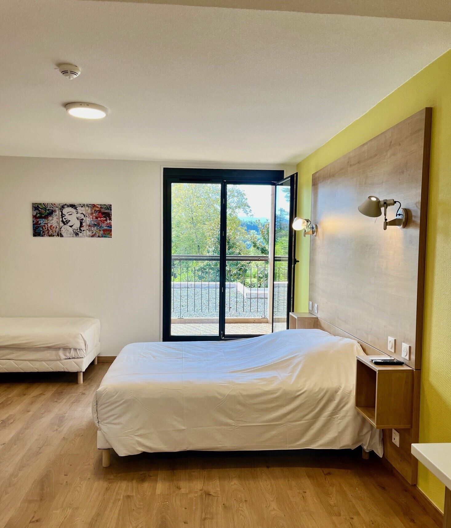 A hotel room with a large bed, a smaller bed, a window with a view of greenery, and a wall-mounted light fixture.